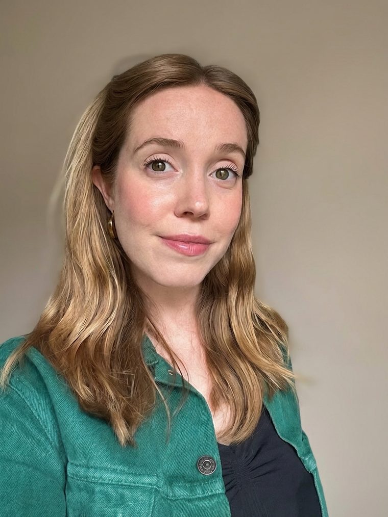 Headshot of Megan Berrisford-Green, a white woman with long wavy blonde hair, smiling warmly at the camera. She is wearing a green denim jacket over a black top and gold hoop earrings, standing against a plain neutral background